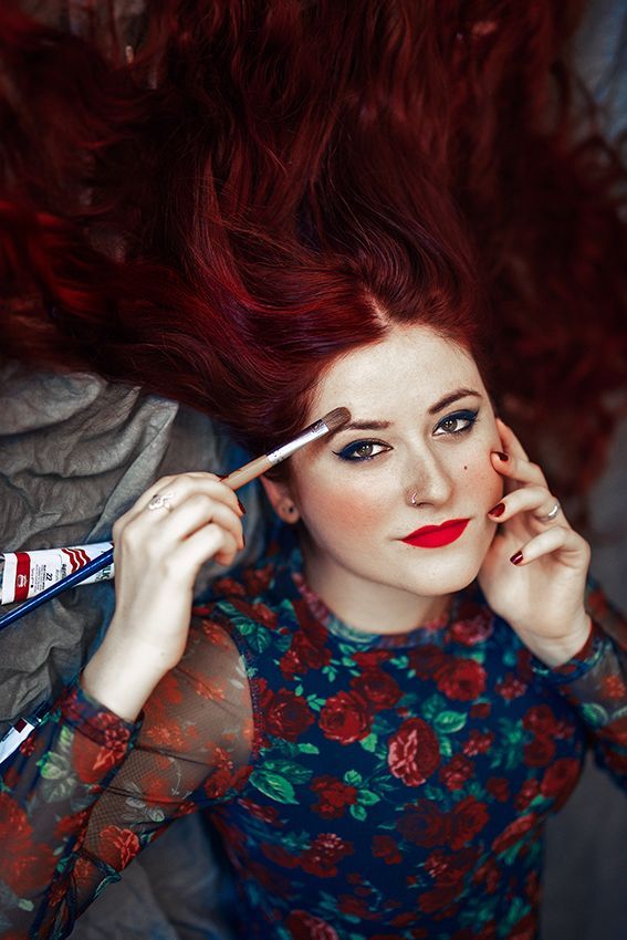 Mujer pintora de pelo rojo largo posando con un pincel dibujando su cara