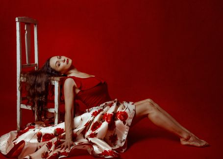 Bailarina posando en el suelo sentada contra una silla con las piernas rectas dobladas y una mano abierta en el suelo con fondo rojo